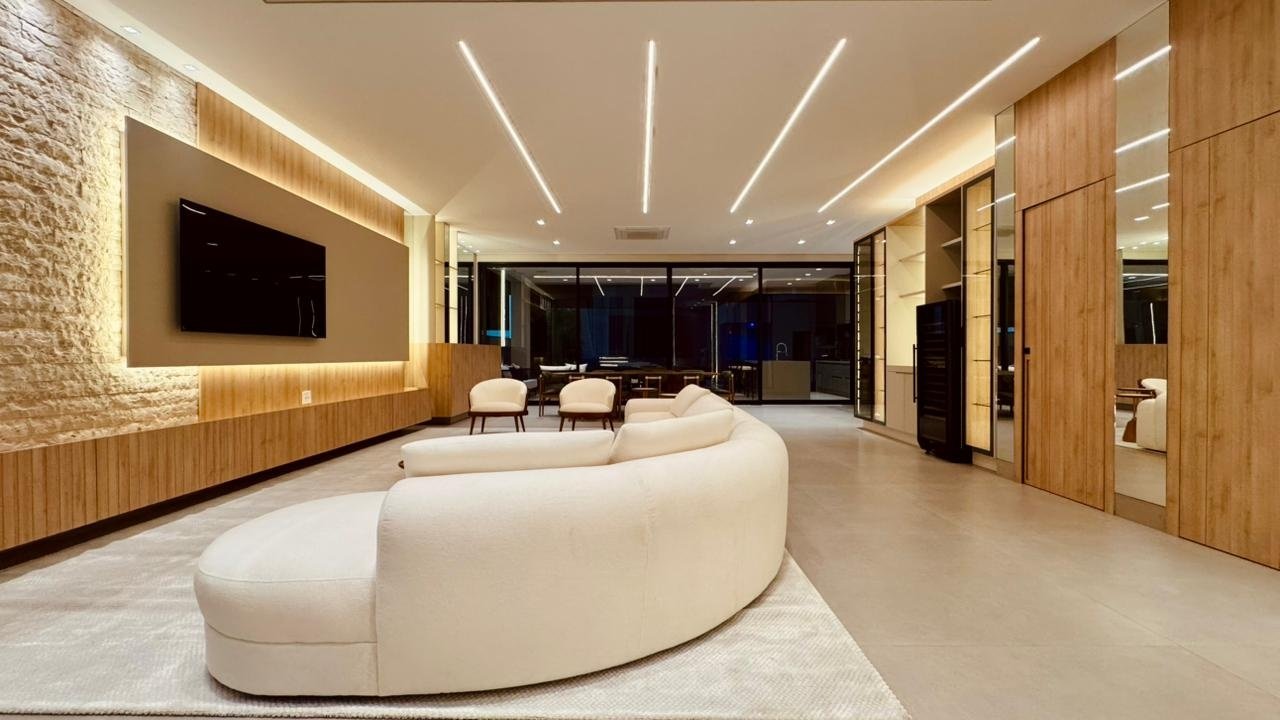 Living da Casa Pinheiro com sofá curvo claro, parede de pedra, painel de TV iluminado e marcenaria em madeira.